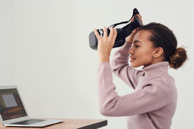 A woman puts on a VR headset in a modern room with a laptop nearby, exploring virtual environments.