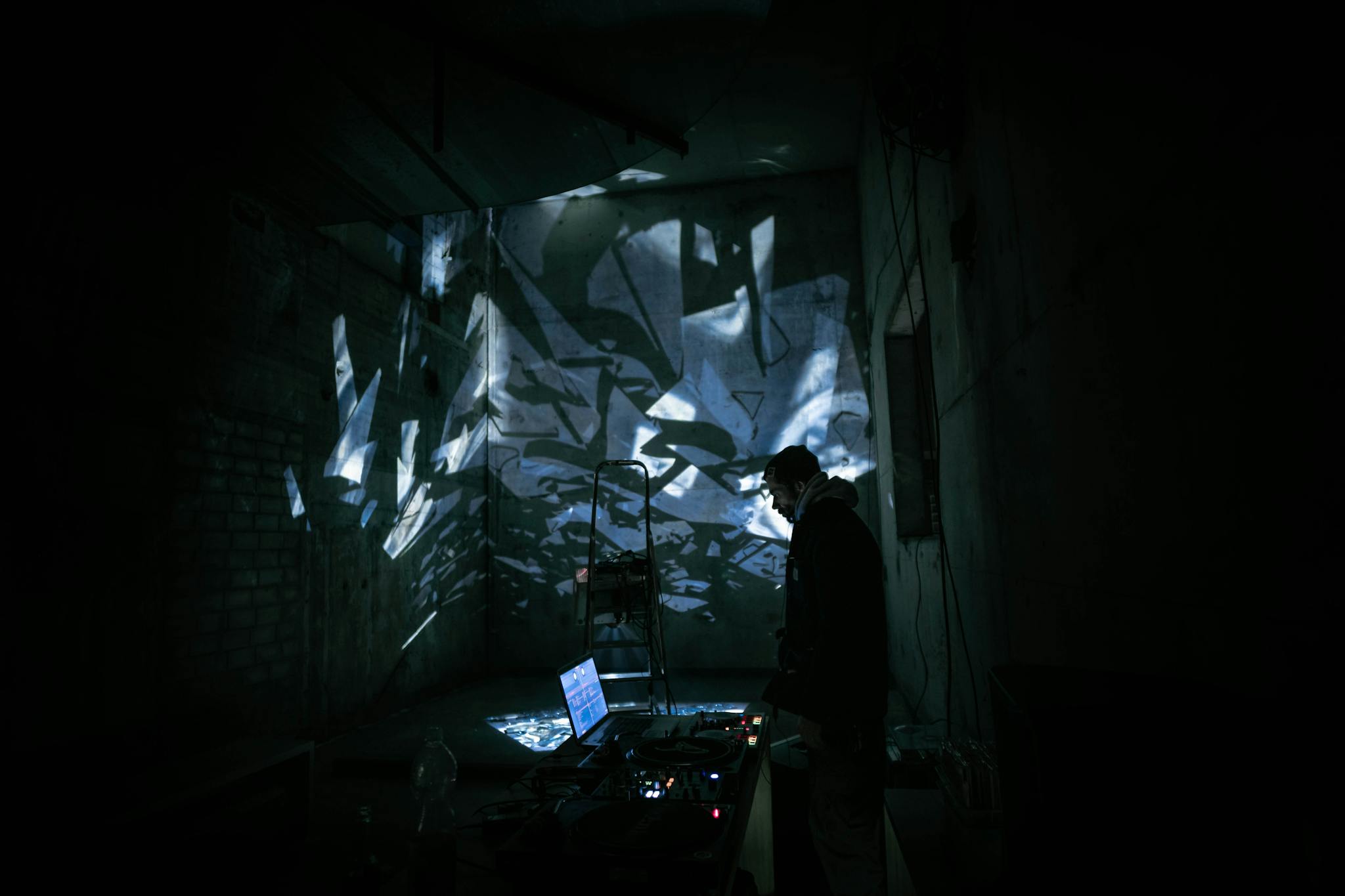A DJ performing in a dark urban setting with abstract light projections creating a moody atmosphere.